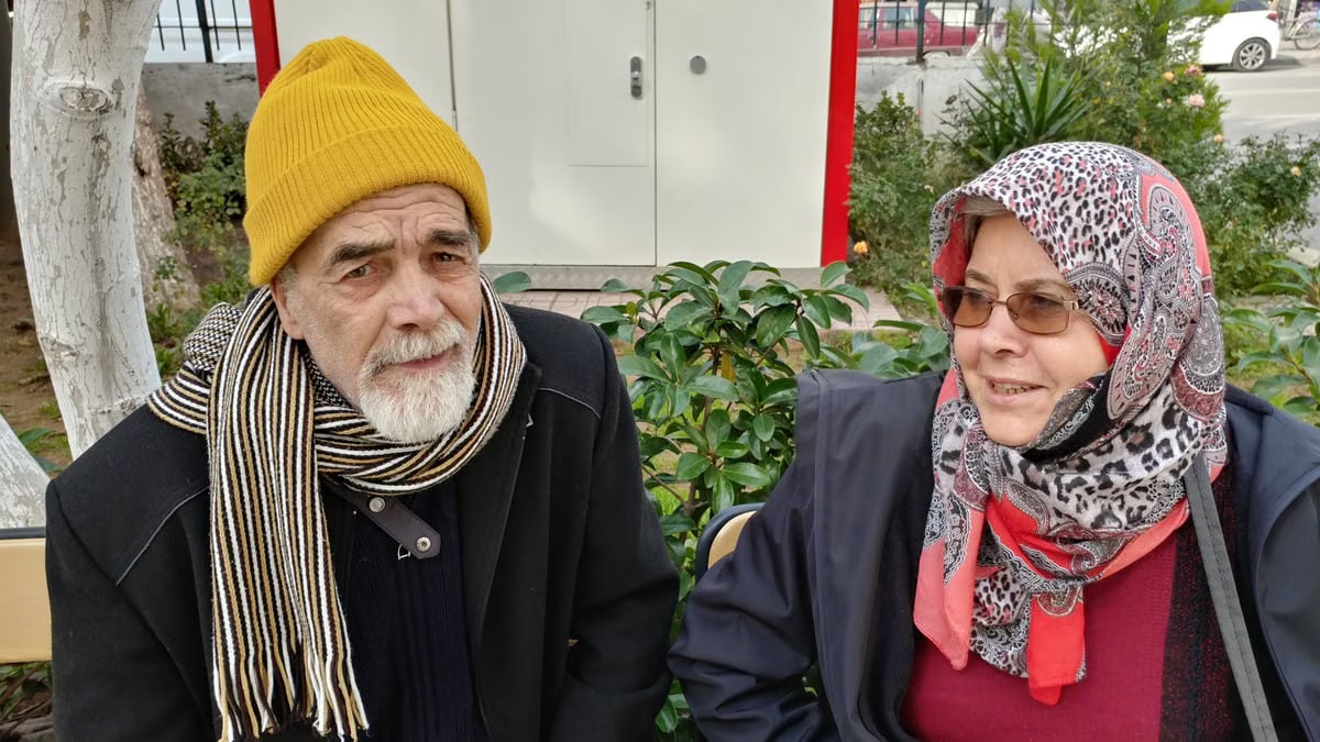 Elderly man and woman sitting outdoors, wearing winter attire with scarves. Bright day scene.